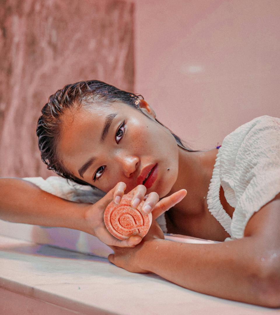 A woman with wet hair leans on a marble surface while holding a circular pink facial soap.