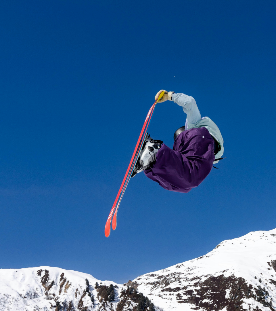 A freestyle skier in purple pants performing an inverted grab against a blue sky and snowy mountains.